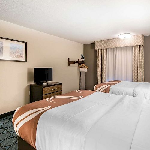 A hotel room with two double beds, a TV on a dresser, a window with curtains, a piece of wall art, and a hanger rack, all in neutral colors.