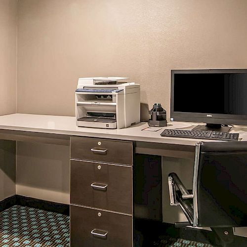 A small office workspace features a computer, a printer, a chair, and a desk with drawers, set against a beige wall and patterned carpet.