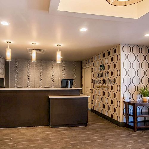 This image shows a hotel reception area with two desks, pendant lights, patterned wallpaper, a small side table with plants, and a vending area.