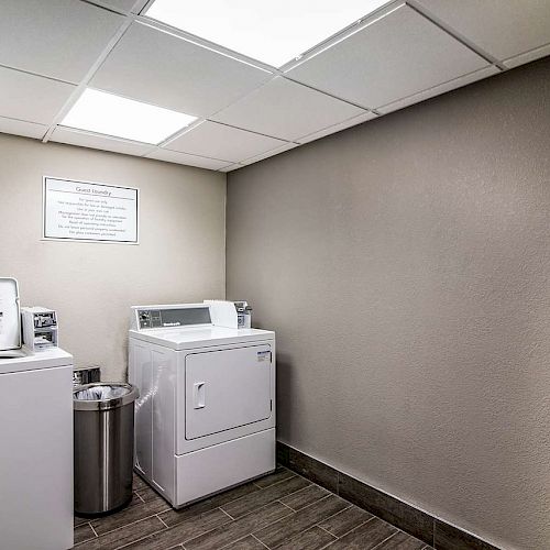 The image shows a laundry room with a washing machine, a dryer, and a trash bin. The room has a tiled floor and a sign on the wall.