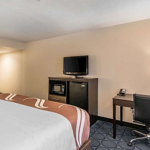 A hotel room with a bed, TV on a dresser, lamp on a desk, an office chair, and a microwave. The room has beige walls and carpeted flooring.