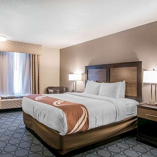 A neatly arranged hotel room featuring a large bed with white linens, two nightstands with lamps, a window with curtains, and a cushioned chair.