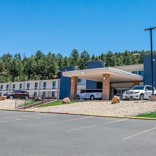 This image shows the exterior of a modern hotel with several parked cars and trees in the background, set against a clear blue sky.