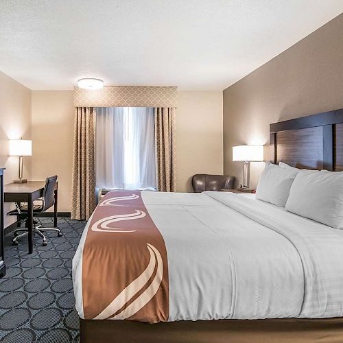 A hotel room with a king-sized bed, desk, chair, TV, and a window with curtains. The room is well-lit and has modern furnishings.