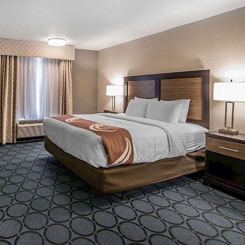 A hotel room with a large bed, two bedside tables with lamps, a TV on a dresser, and a window with curtains. The room has patterned carpet flooring.