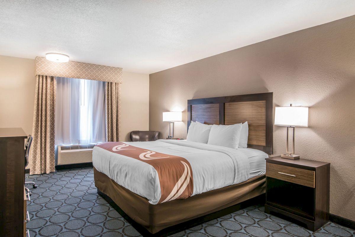 A hotel room with a large bed, two nightstands with lamps, a window with curtains, a chair, and a dresser in a neat, well-lit setting.