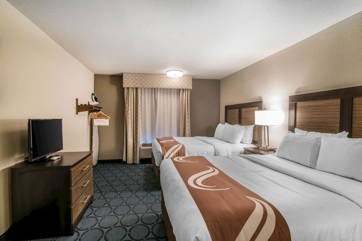 A modern hotel room with two double beds, a flat-screen TV, a window with curtains, a dresser, and a lamp on a nightstand between the beds.