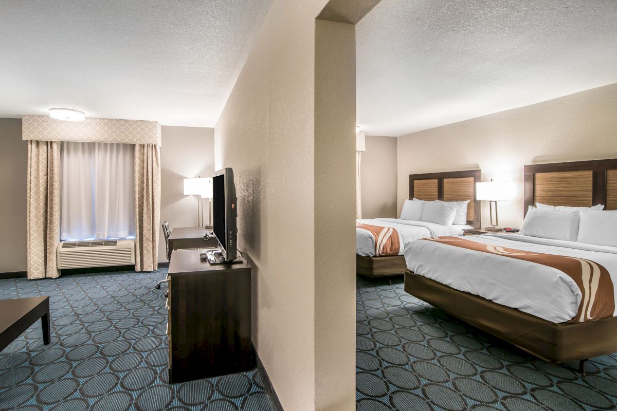 The image shows a hotel room divided by a partial wall, with a TV, seating area, two beds, bedside tables and lamps.