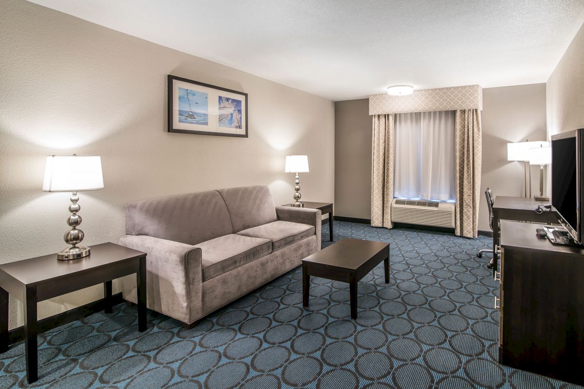 A neatly arranged hotel room with a sofa, center table, two lamps, a desk with a chair, a TV, and wall art, all illuminated by soft lighting.