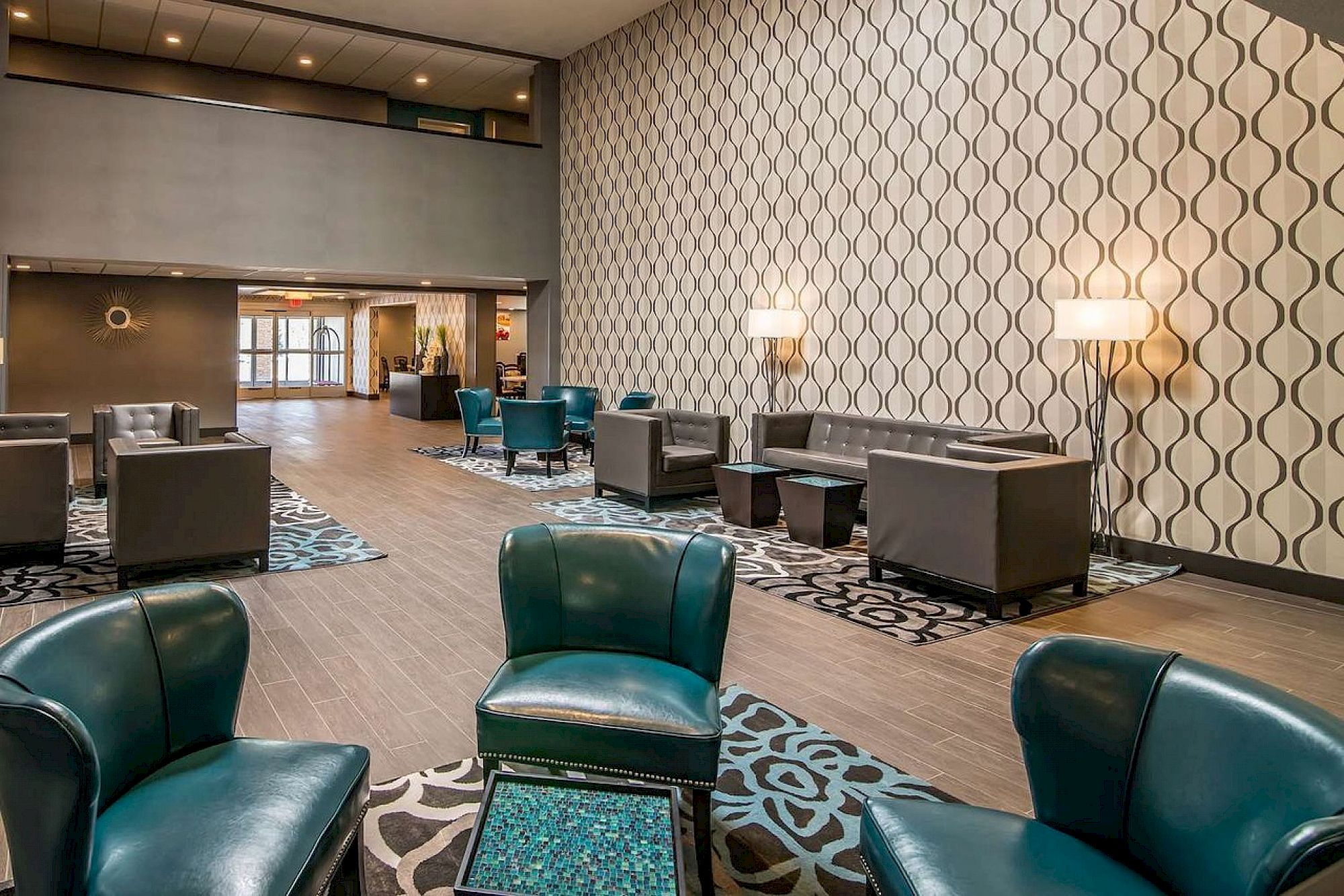 A modern lounge area with patterned wallpaper, teal leather seating, tables, and lamps, featuring a mix of wood and carpet flooring.