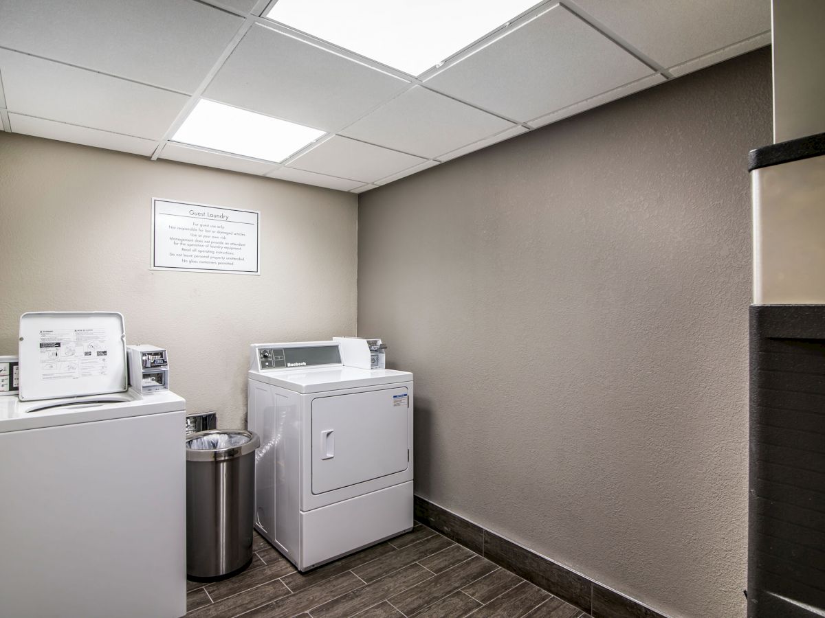 The image shows a small laundry room with a washing machine, dryer, and a metal trash can, all against a beige wall under fluorescent lighting.