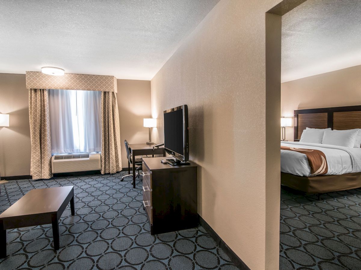 This image shows a hotel room with a seating area, a TV, a desk, and a bedroom with a bed in the background.