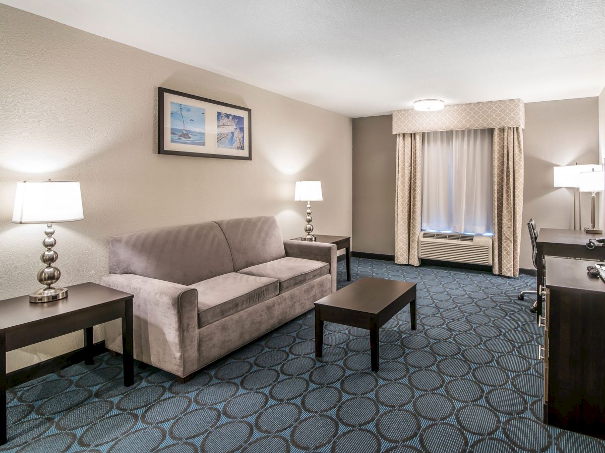 The image shows a hotel room with a sofa, two lamps, a coffee table, a TV on a cabinet, a desk, and a window with curtains.