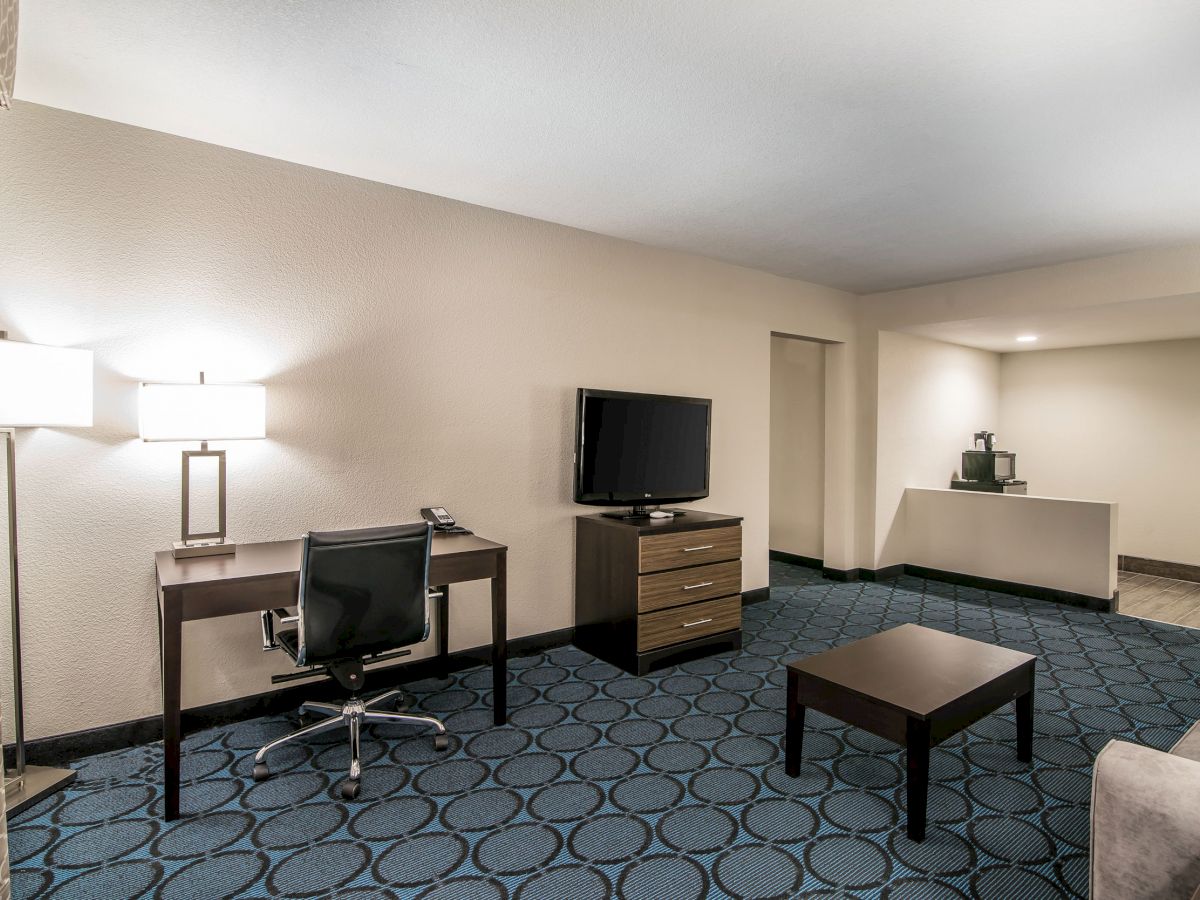 The image shows a hotel room with a desk, chair, TV, drawer, coffee table, lamp, and a coffee maker on a countertop.