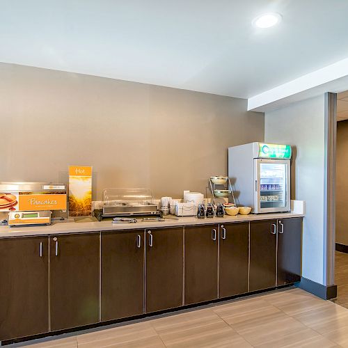 This image shows a hotel breakfast area with a counter containing a juice dispenser, cereal, a mini fridge, coffee machine, and pancake maker.