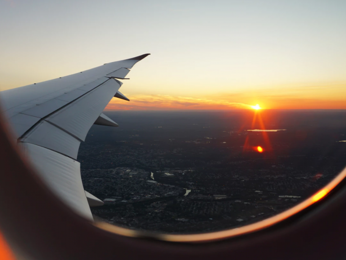 View from an airplane window showing a wing and a sunset over a landscape below. The sky is beautifully lit with warm, golden hues.