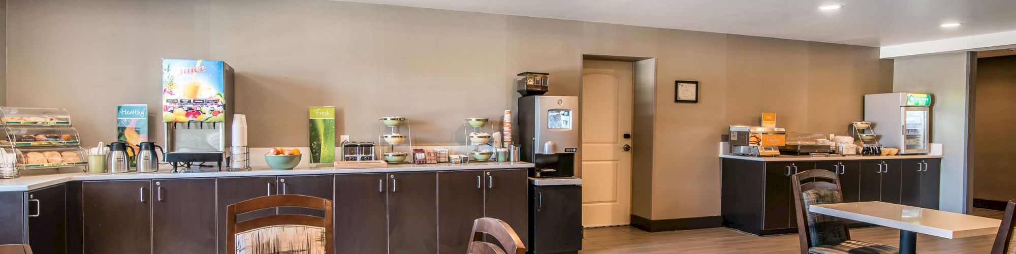 Image shows a hotel breakfast area with various food and beverage stations along the wall and tables and chairs for seating.