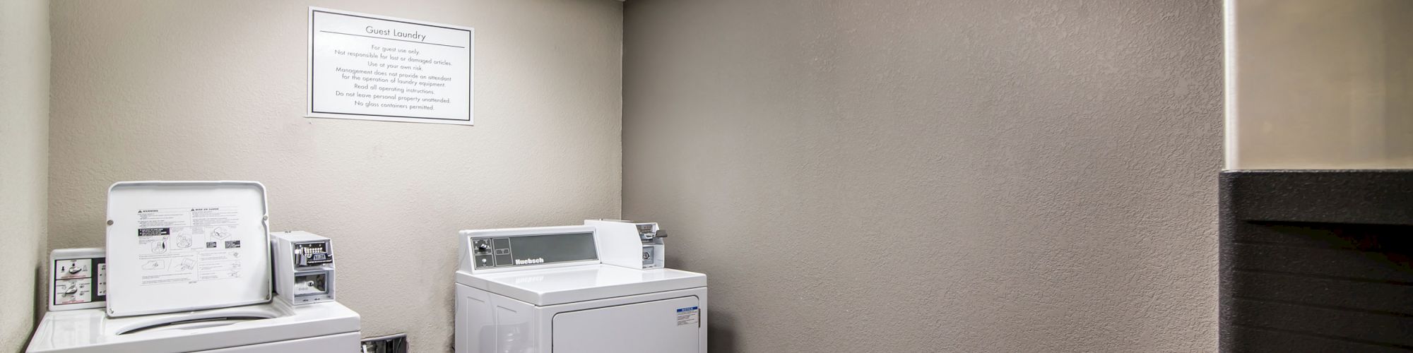 The image shows a small laundry room with a washing machine, a dryer, a trash can, and a sign on the wall, all under a well-lit ceiling.