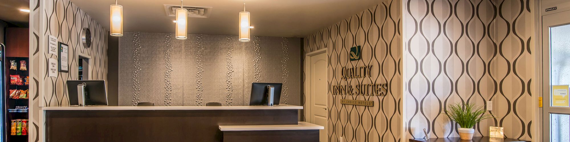 This image shows a modern, well-lit hotel reception area with a front desk, decorative wall pattern, and a small table with plants and decor items.