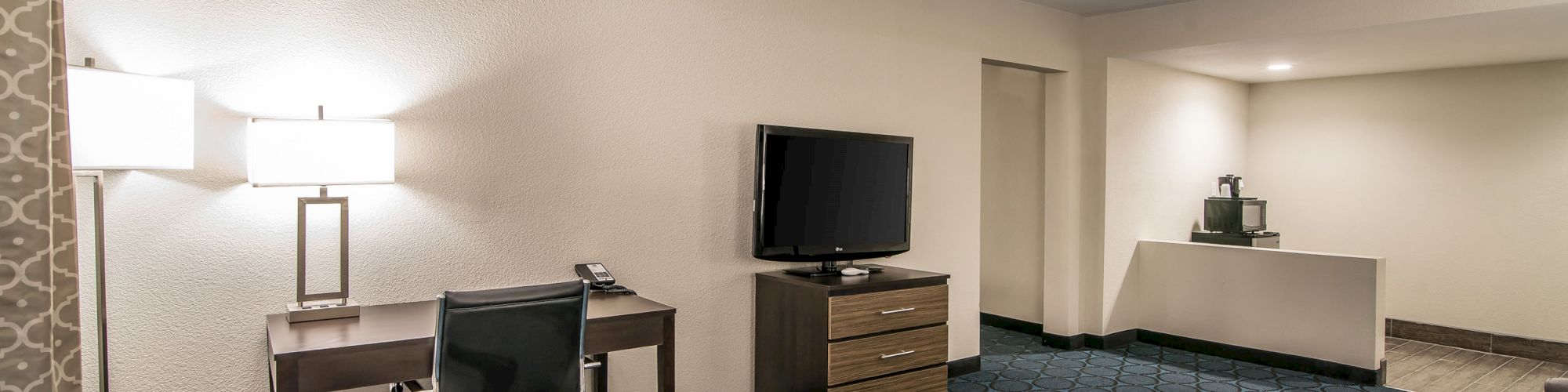 A hotel room with a desk, chair, lamp, flat-screen TV on a drawer, coffee maker, and a small table on a patterned blue carpet floor.